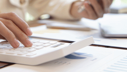 A hand using a calculator at a desk with paper bills.